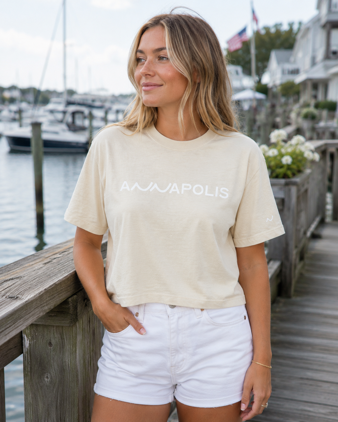 A woman standing on a wooden dock by the water wearing a cream-colored cropped t-shirt with 'ANNAPOLIS' printed in white across the chest, paired with white denim shorts.