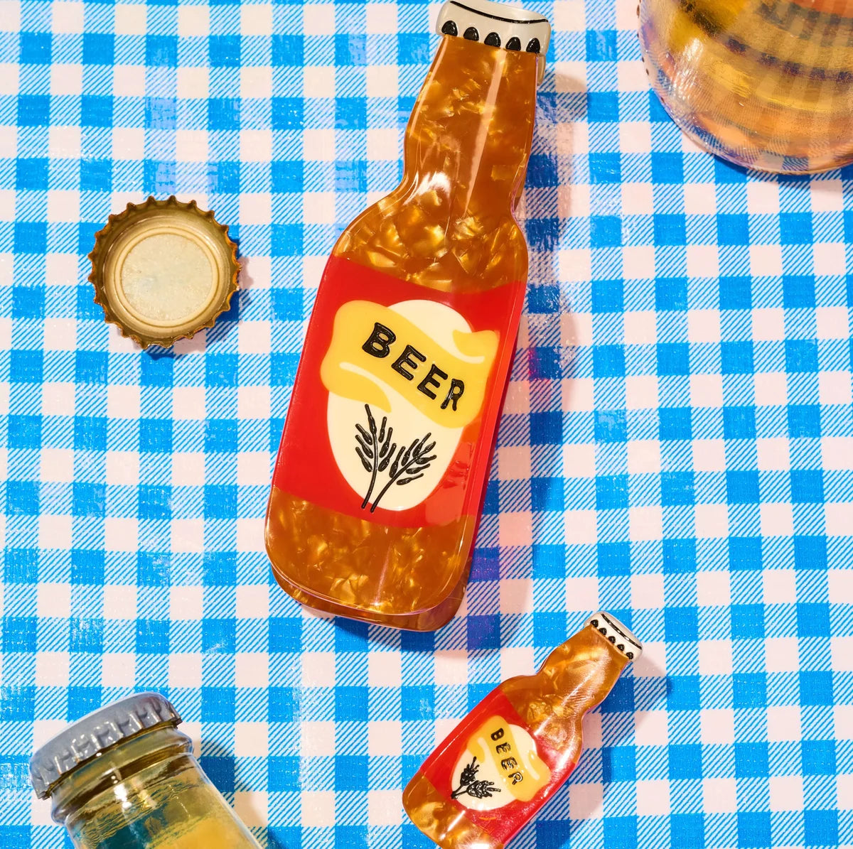 Large and mini beer bottle hair clips with amber pearlescent details on a blue-and-white gingham background. West Annapolis Oktoberfest.